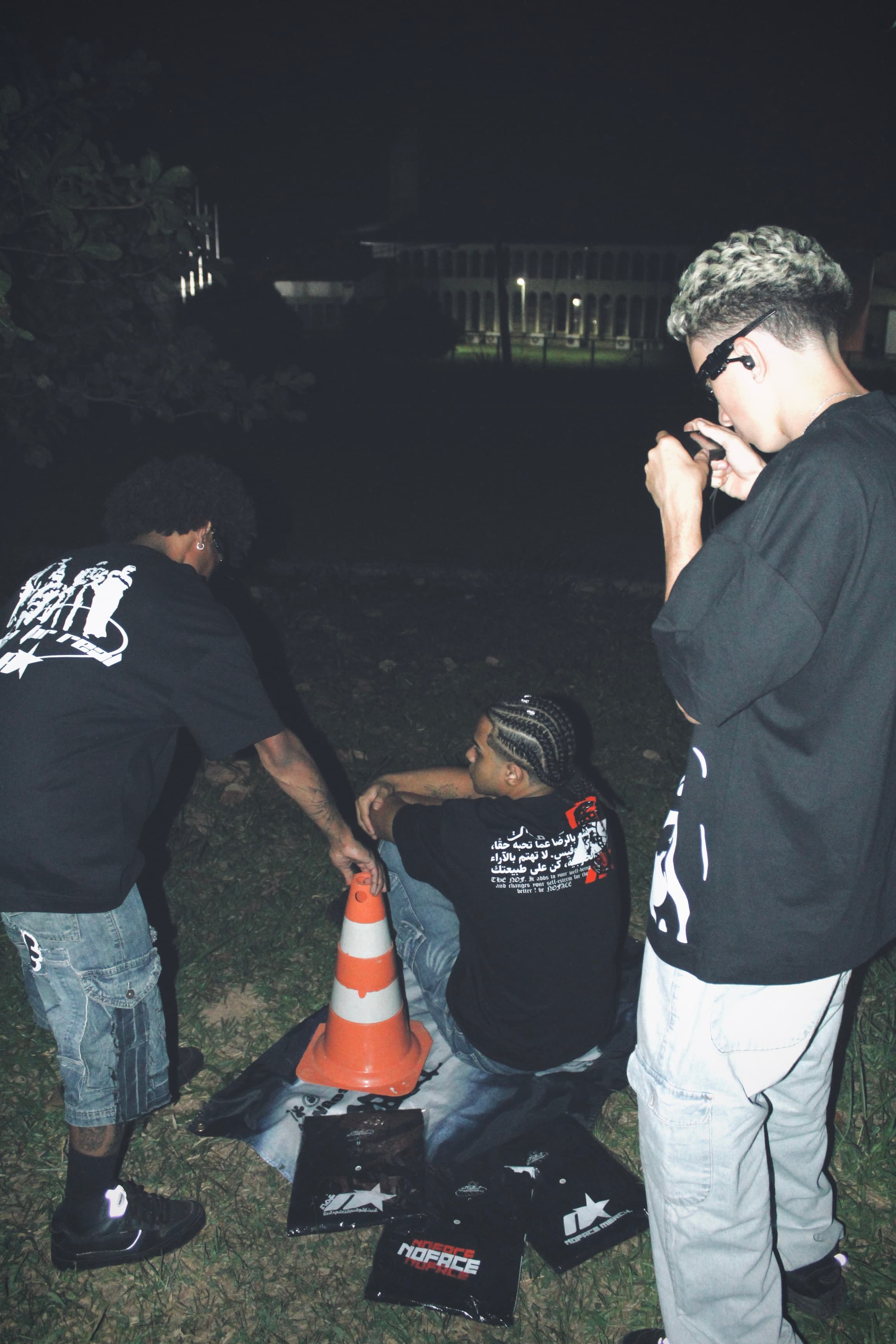 tres homens em um gramado no meio das camisetas da marca 'NOFACE' com um cone ao lado, um deles esta tirando foto dos outros