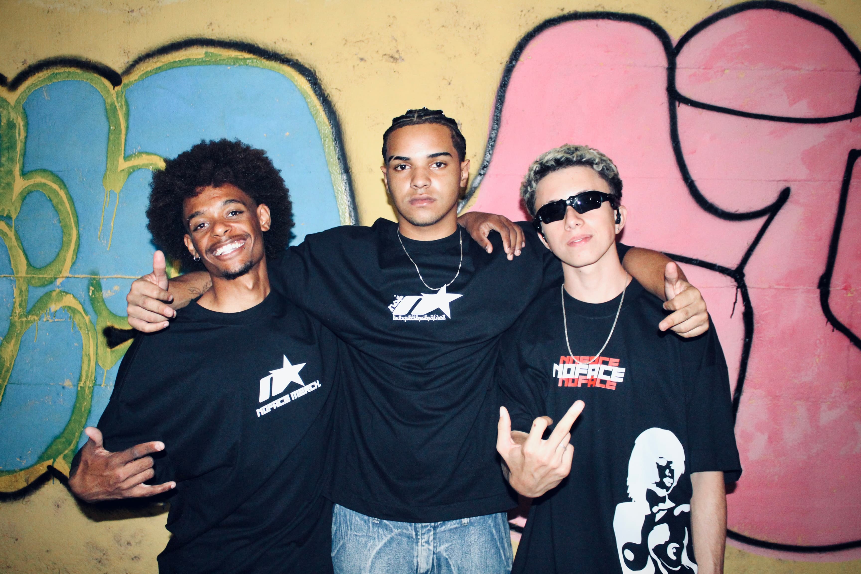 tres homens abracados fazendo pose para a camera, na frente de uma parede com pichação