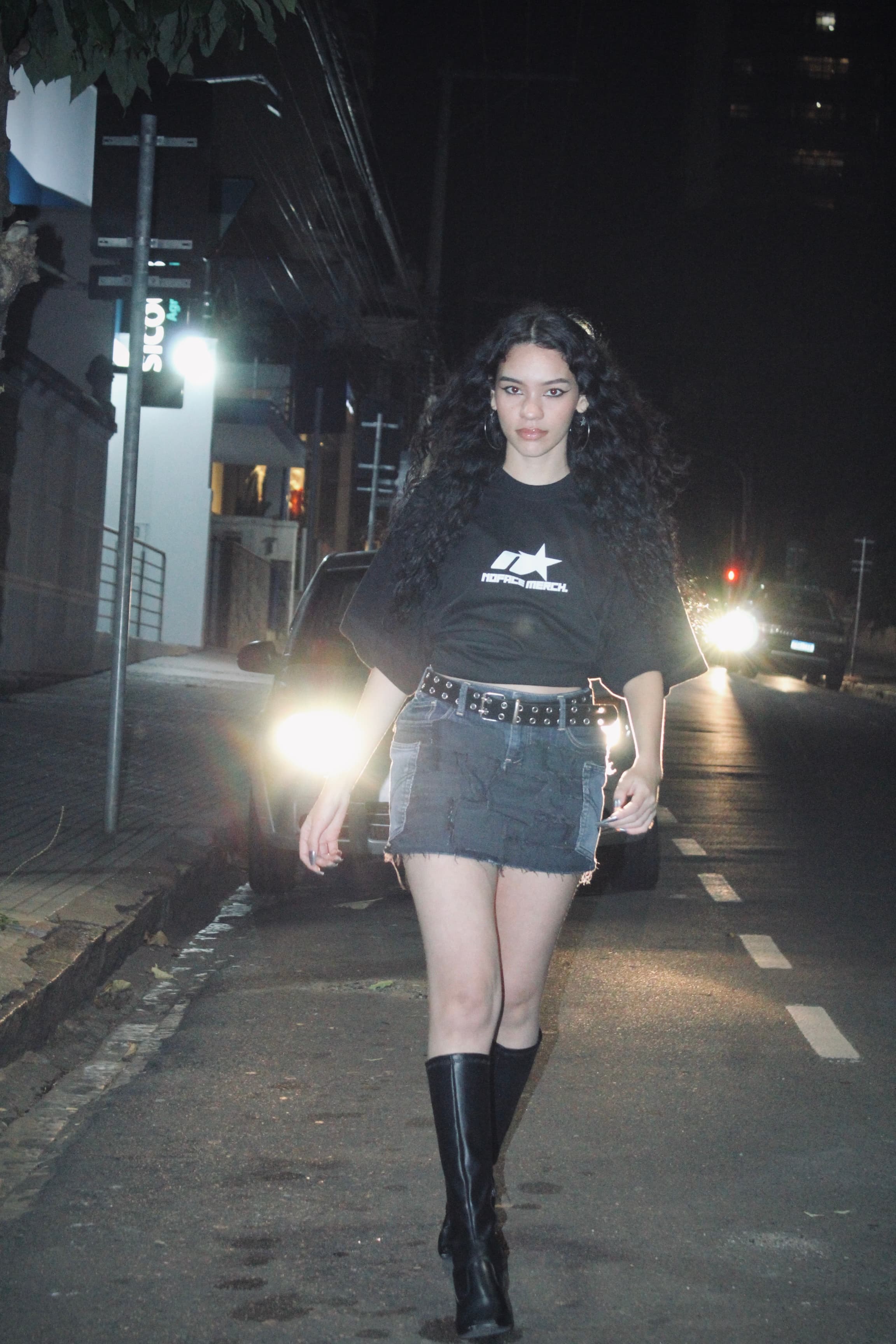 mulher andando na frente do farol de um carro usando a camiseta escrito 'NOFACE MERCH' com o logo da marca 'NOFACE'